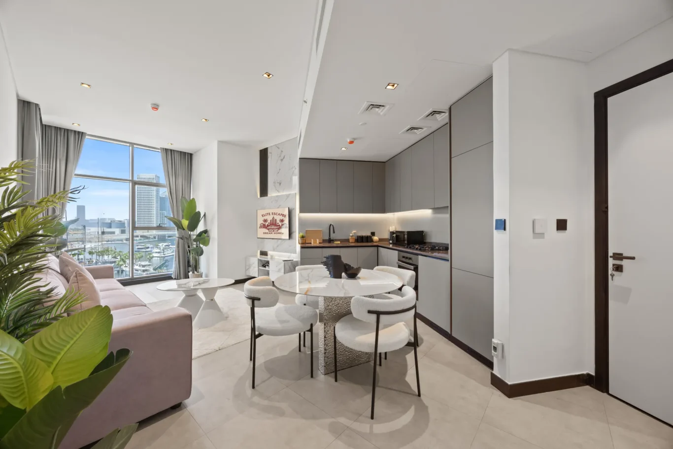 Open-plan living area with a pink sofa, round marble tables, white chairs, and a gray kitchen, city skyline visible through large windows.