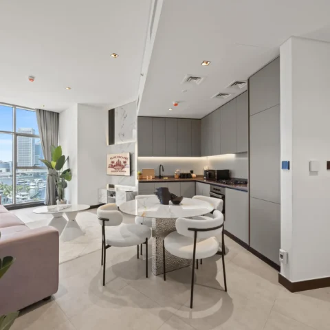 Open-plan living area with a pink sofa, round marble tables, white chairs, and a gray kitchen, city skyline visible through large windows.