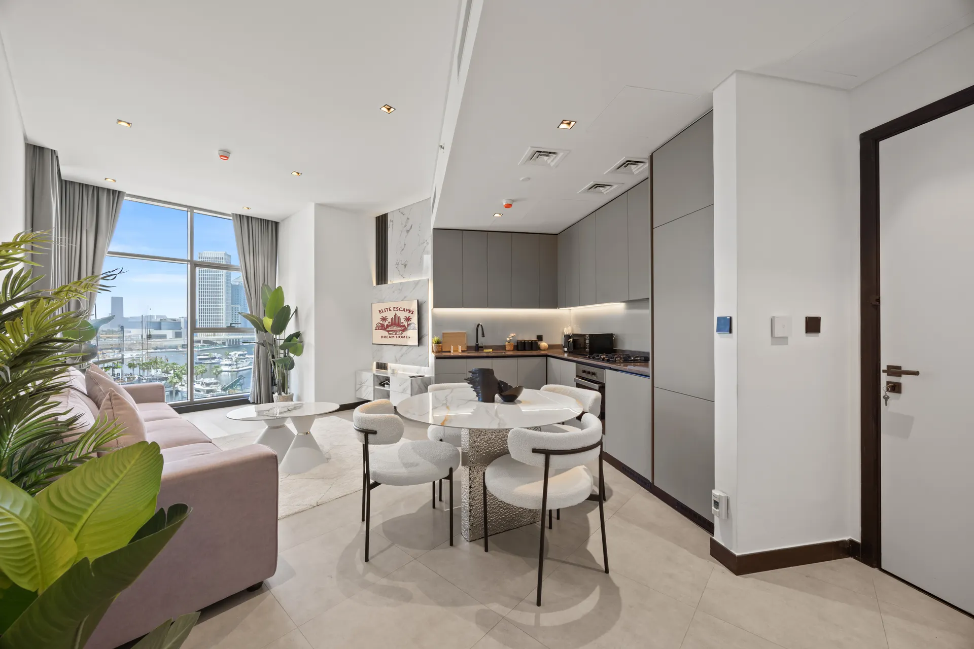 Open-plan living area with a pink sofa, round marble tables, white chairs, and a gray kitchen, city skyline visible through large windows.