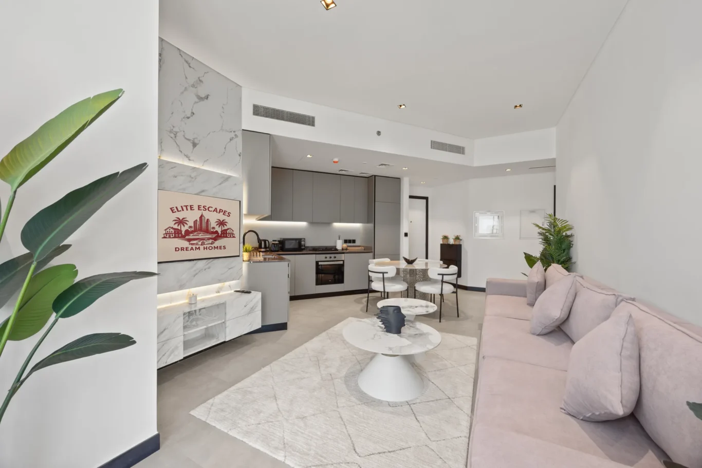 Open-plan living room and kitchen with a pink sofa, white marble accents, and greenery; sign reads 'Elite Escapes Dream Homes'.