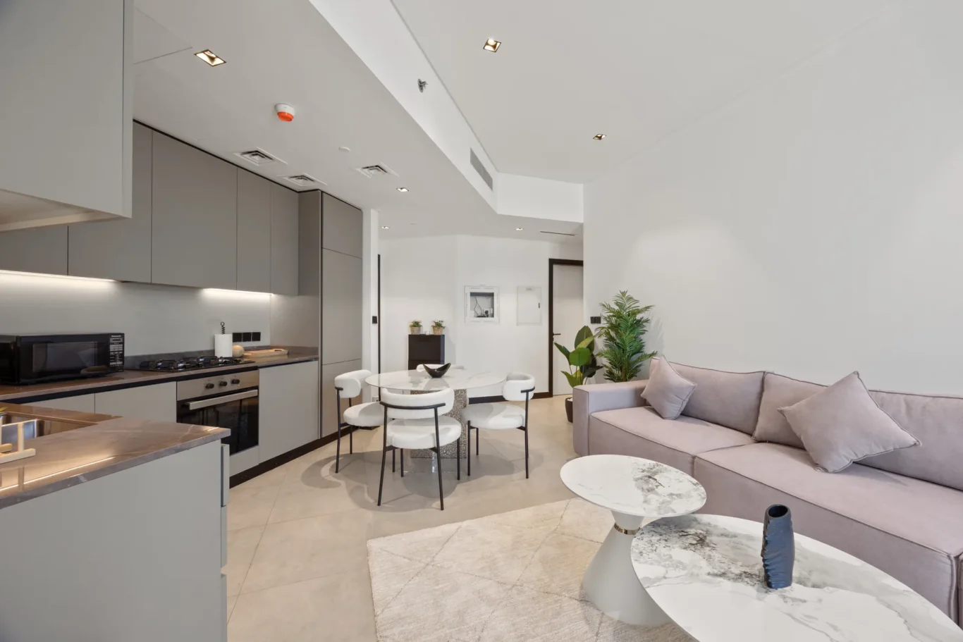Open-plan kitchen and living area with gray cabinetry, breakfast island, round marble tables, and a light beige sofa with cushions beneath bright recessed lighting.