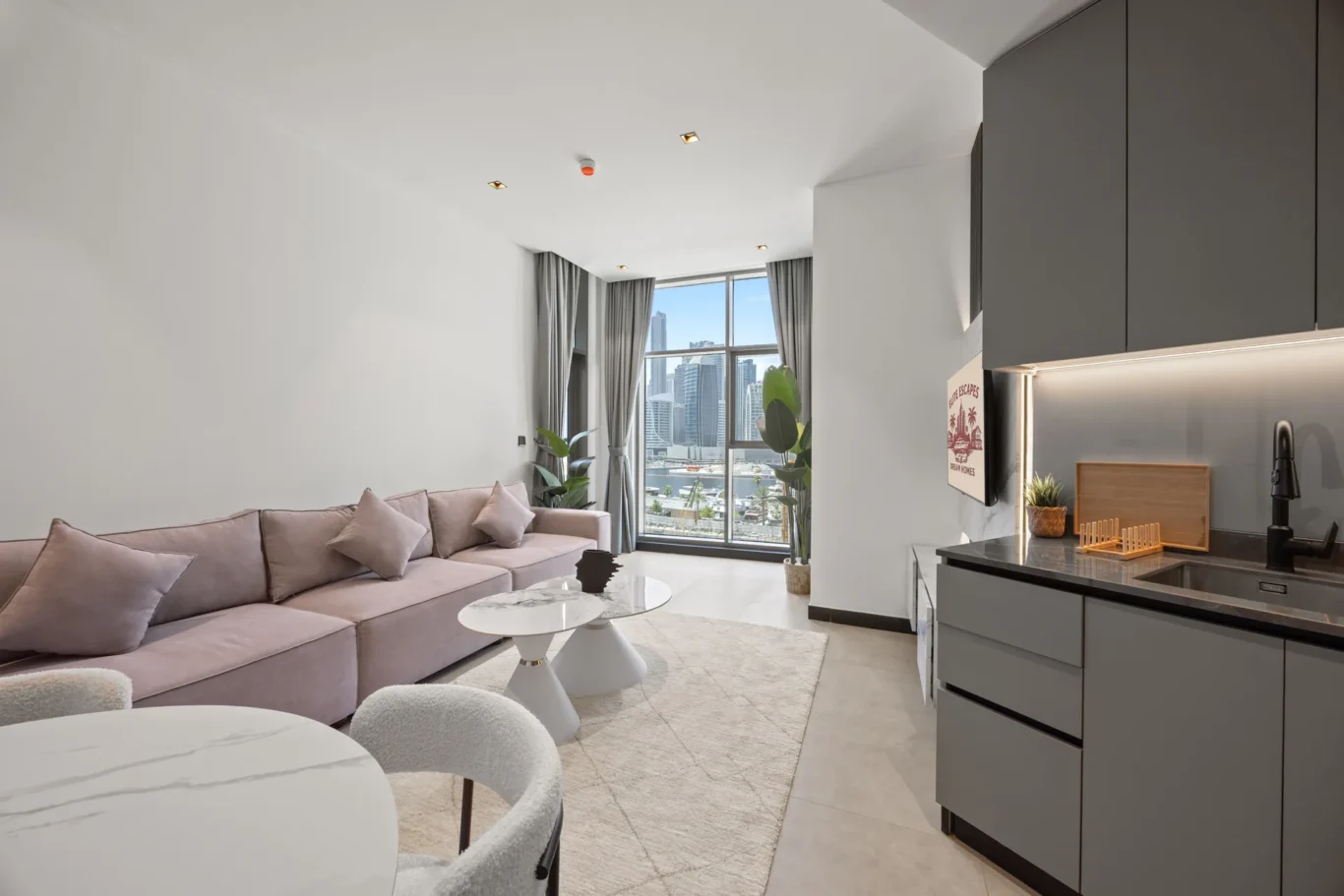 Open-concept living area with a pink sectional sofa, white marble-topped tables, and large windows overlooking a city skyline.