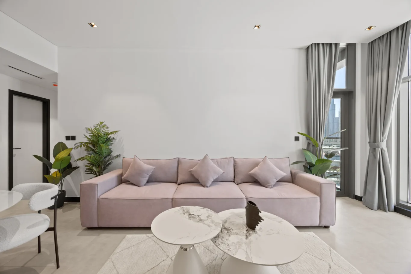 Modern living room with a pale pink sofa, round marble coffee tables, and potted plants by large windows with gray curtains.