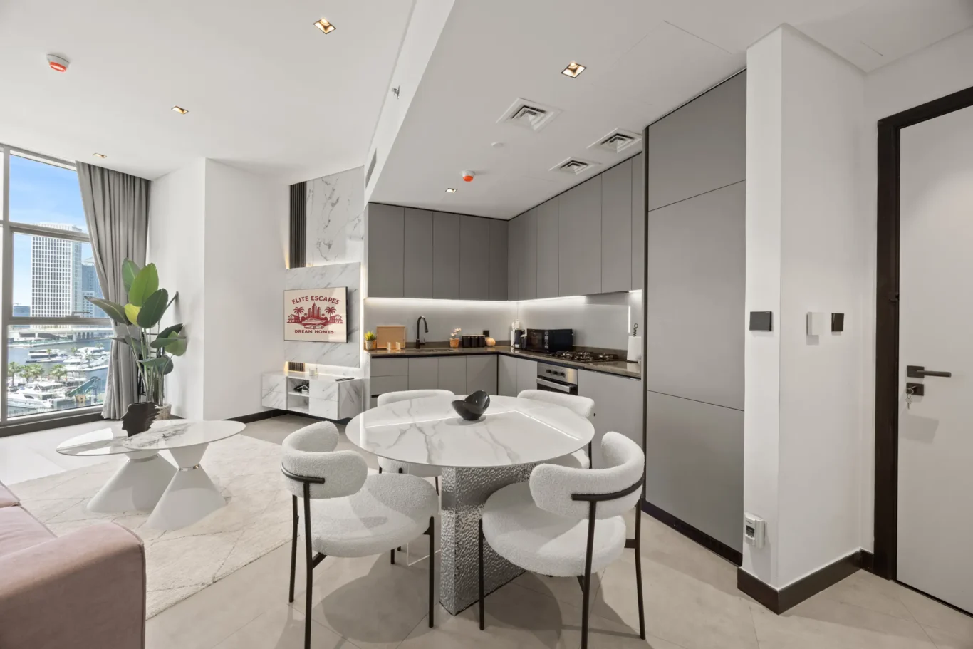 Open-plan kitchen and dining area with gray flat-panel cabinets, marble-topped island, and round marble tables surrounded by white cushioned chairs; large window shows a city skyline and water.”] ,
