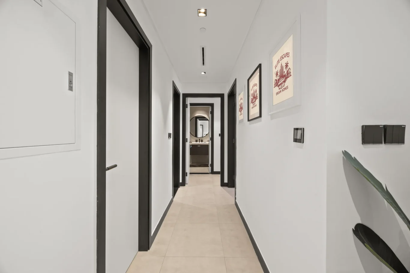 Long white hallway with black door frames and framed art along the right wall, leading to a round-mirrored bathroom at the end.