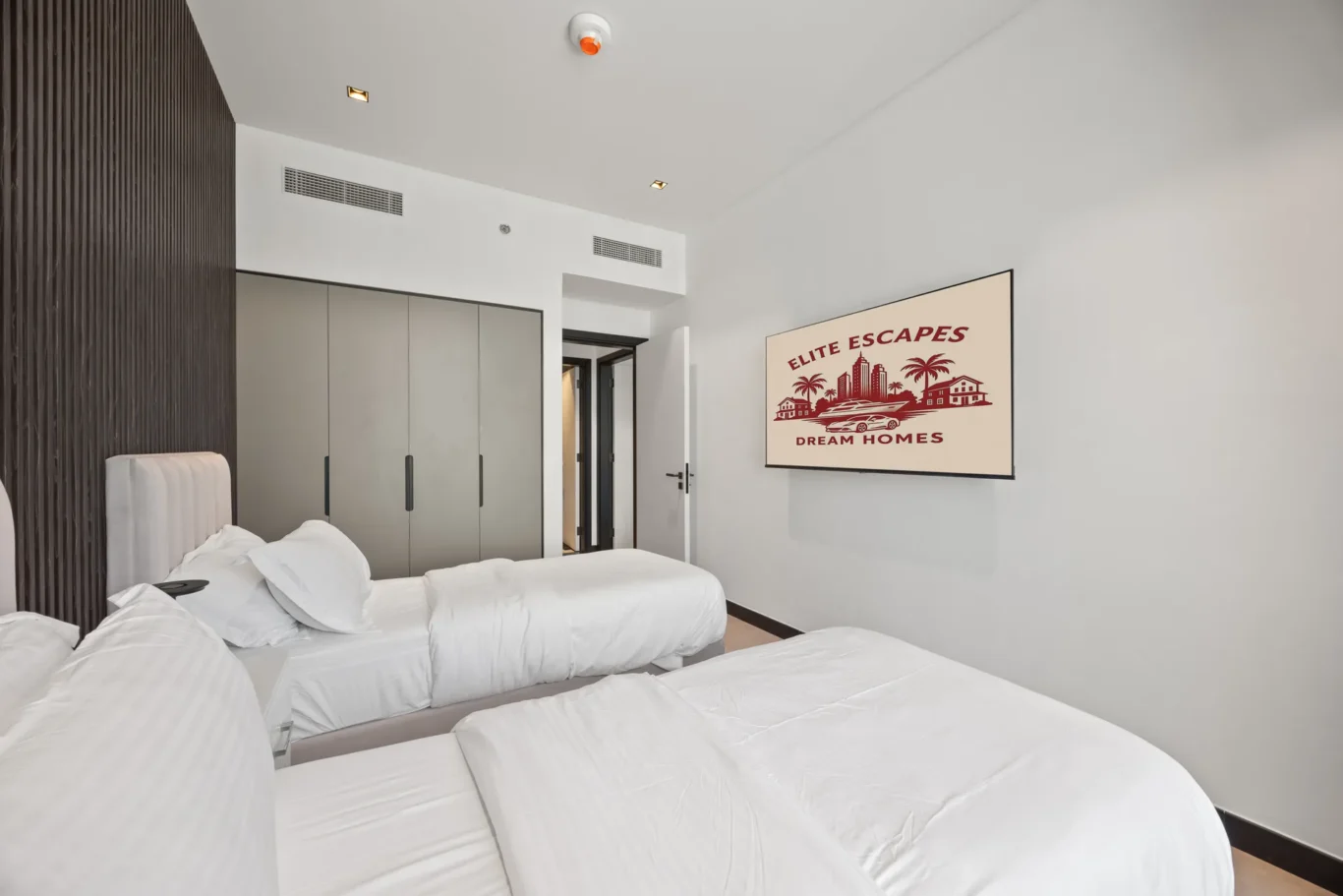 Hotel room with four single beds in white linens, a dark wood paneled accent wall, gray wardrobe cabinets, and a framed mural reading 'Elite Escapes Dream Homes'