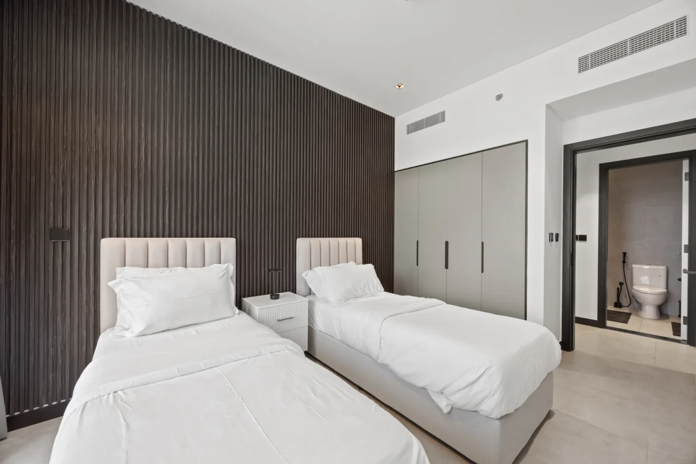 Twin beds in a modern hotel room with a vertical dark wood panel wall and an open bathroom door to a toilet area on the right.