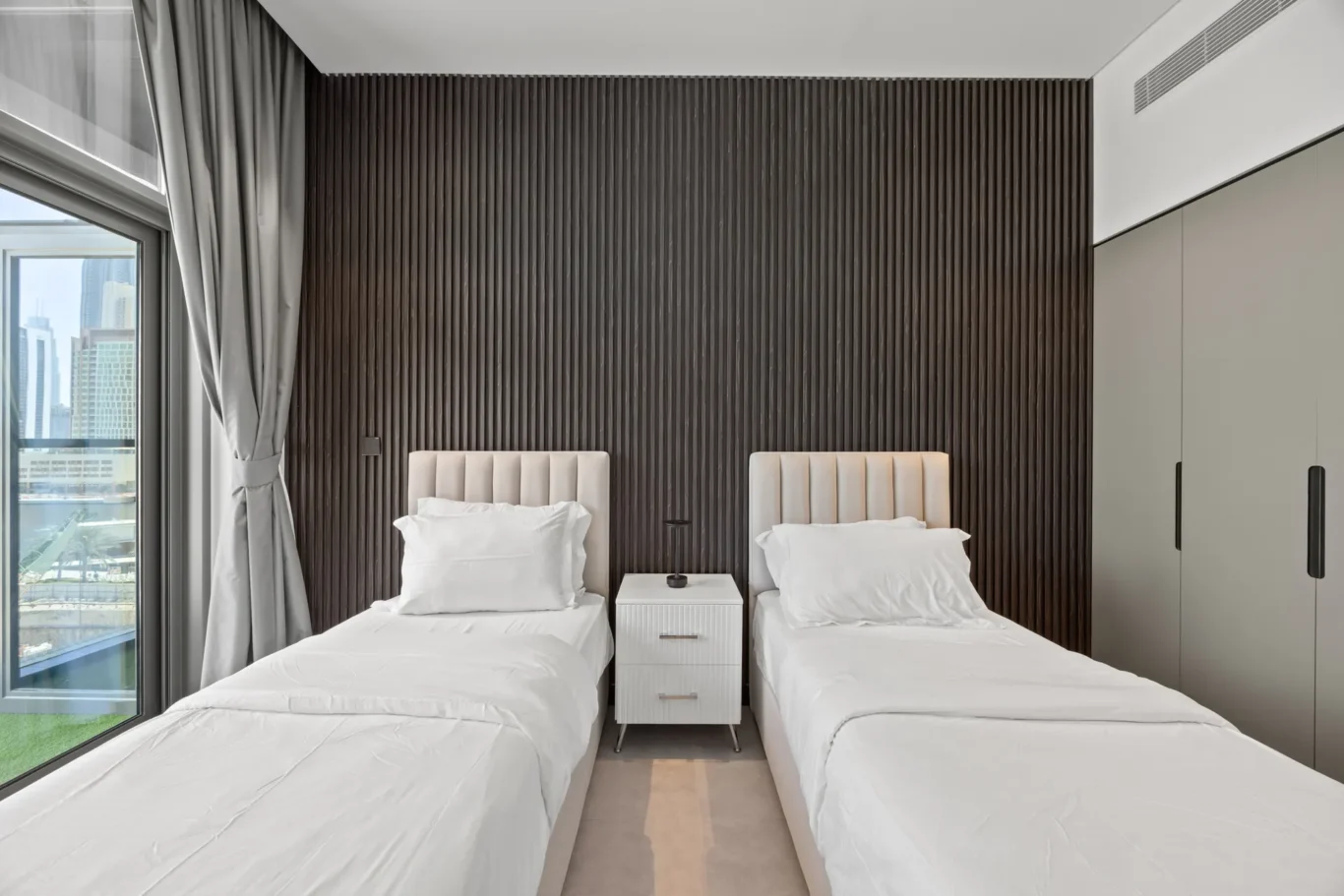 Twin hotel beds with white linens and a central nightstand against a dark vertical wood panel wall.