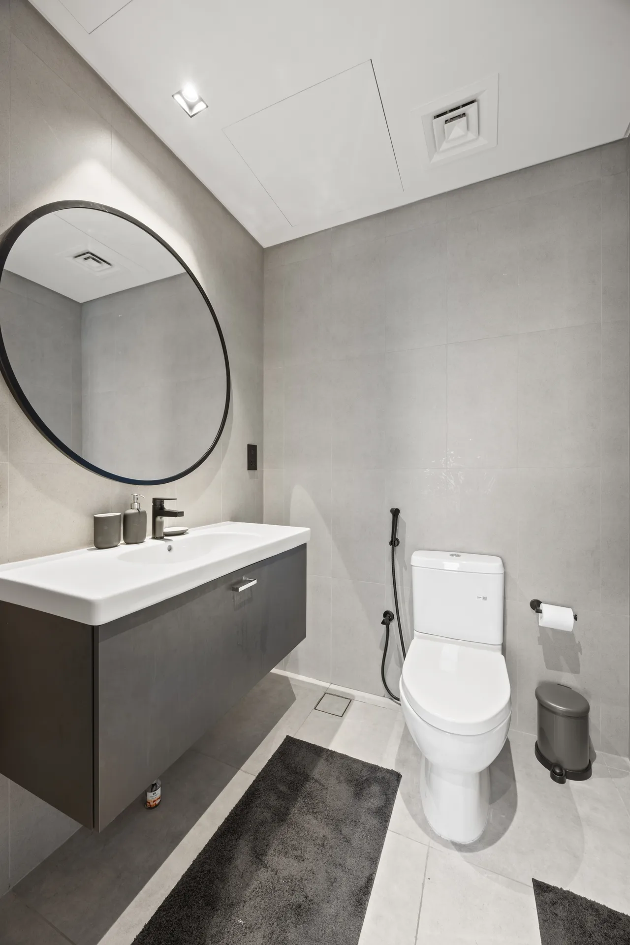 Modern gray bathroom with a round black-framed mirror above a white sink and dark vanity, next to a toilet and bidet spray.