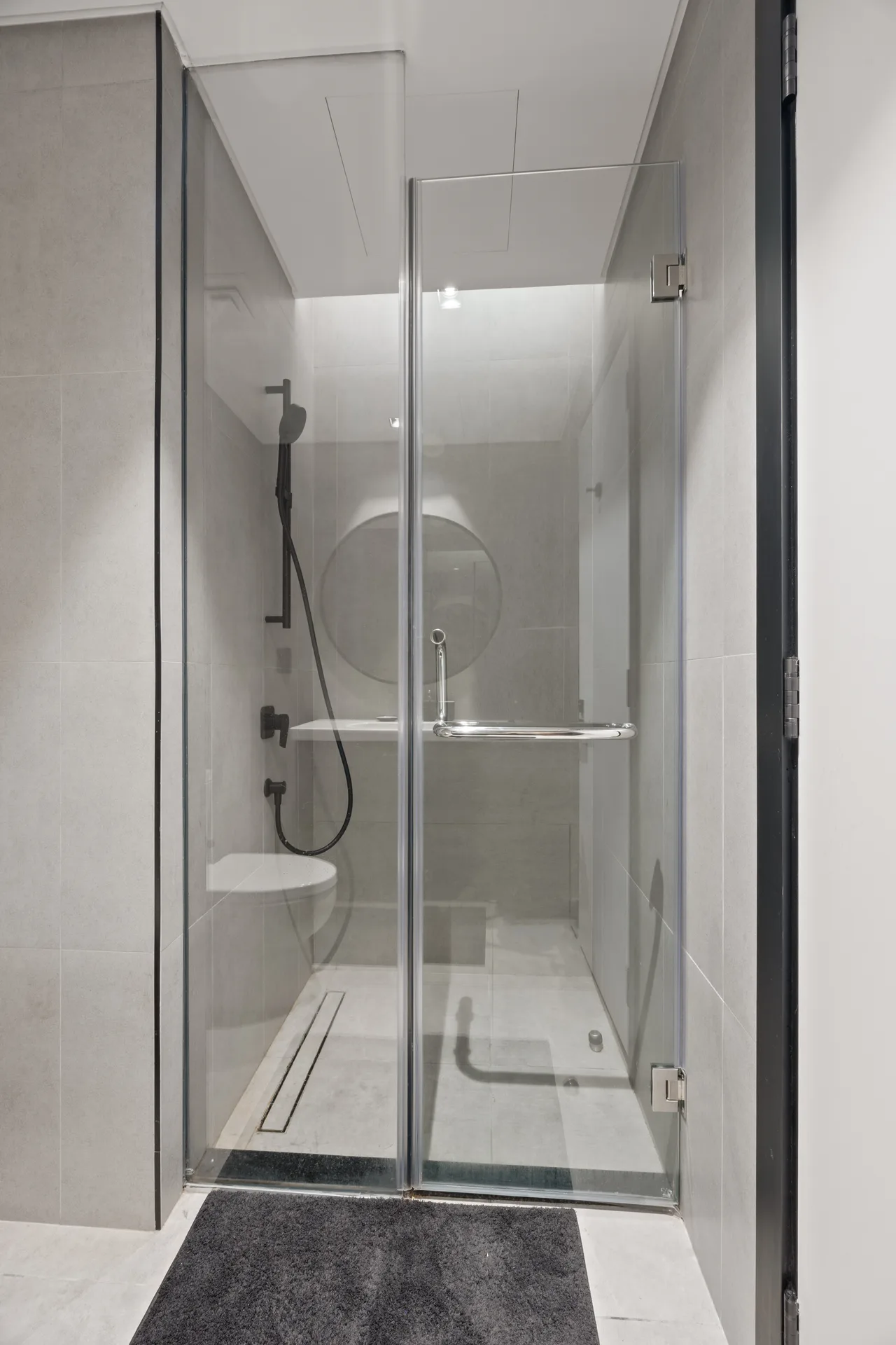 Glass-walled shower stall with a black handheld shower, chrome bars, and gray tiled walls inside the bathroom.