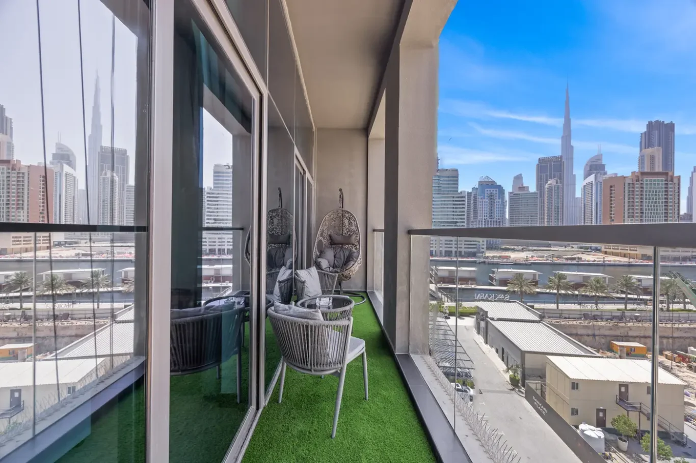 Balcony with artificial grass, gray wicker chairs, and hanging egg chairs, overlooking a modern city skyline.