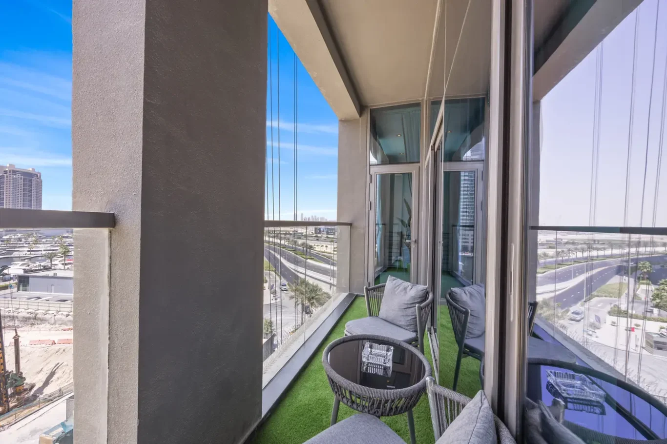 Balcony seating area with gray cushions, a round glass table, and artificial grass, overlooking a city highway and blue sky.