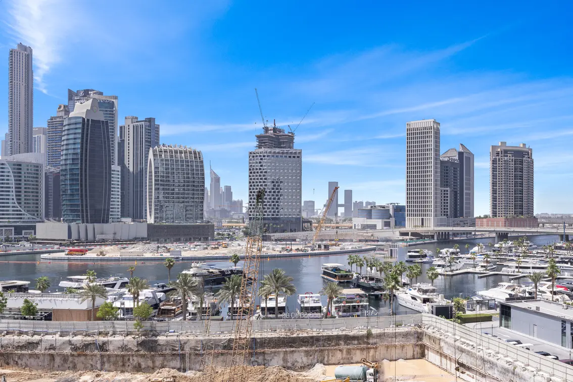 Skyline of modern skyscrapers by a marina with yachts under a bright blue sky, cranes visible in the foreground.