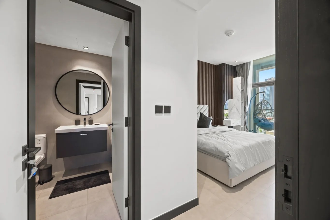 Open doorway into a modern hotel suite: left shows a bathroom with a round black-framed mirror above a white sink; right reveals a bedroom with a large bed and a wooden slat wall near a window.