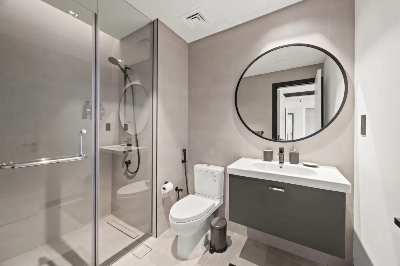Modern grey bathroom with a glass shower enclosure, round mirror, and white vanity cabinet beneath the sink.