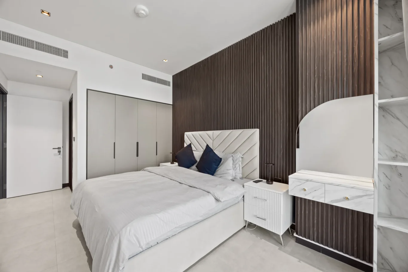 Modern bedroom with a gray upholstered bed, dark wooden slat accent wall, and marble vanity. The space looks bright and contemporary with clean lines.