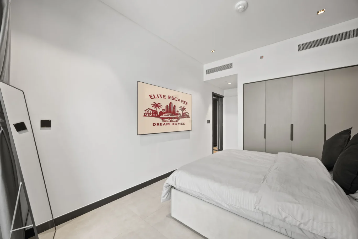 Hotel-style bedroom with a wall-mounted artwork reading 'Elite Escapes Dream Homes' over a bed area. Arms-length mirror leans against the wall; large gray wardrobe on the right.