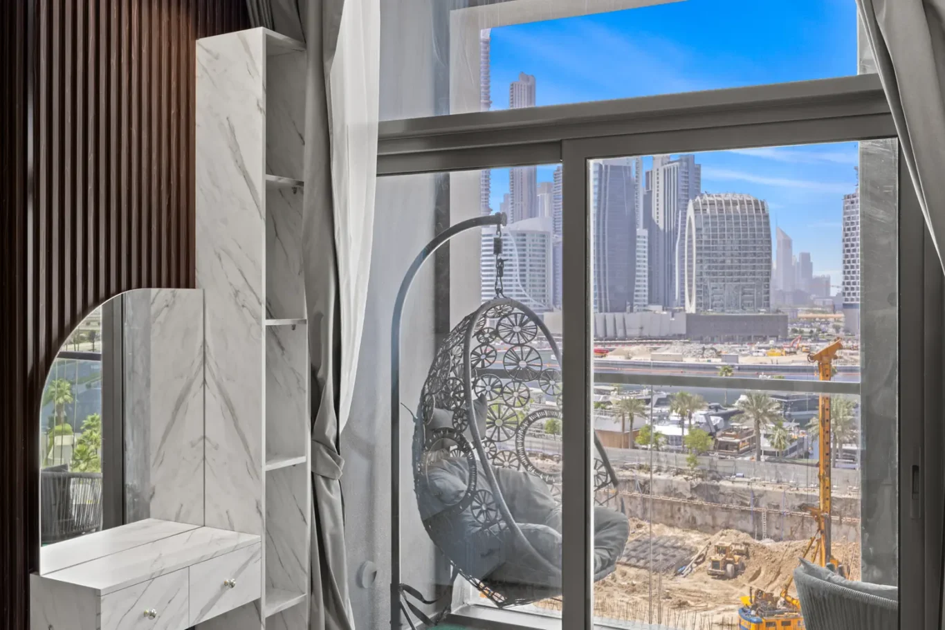 Marble-topped vanity and tall marble shelving unit inside a modern room, with a curved metal-framed hanging chair by a large window overlooking a construction site and city skyscrapers.