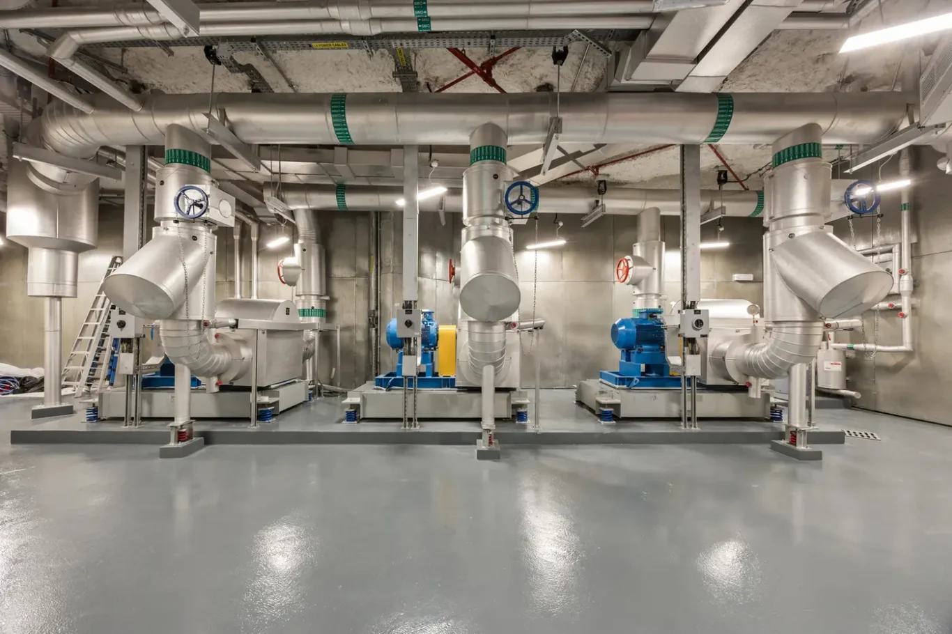 Industrial mechanical room with large stainless steel ducts, valves, and pumps arranged in a row for fluid handling or HVAC systems.