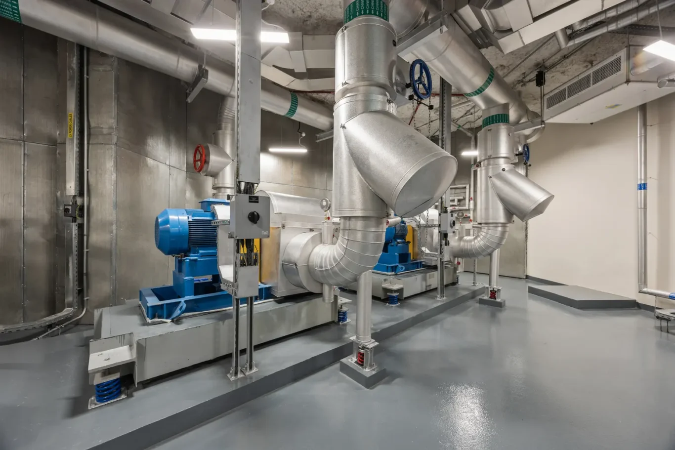 Industrial mechanical room with large galvanized ducts, blue motors, and extensive metal piping forming a high-capacity ventilation system.