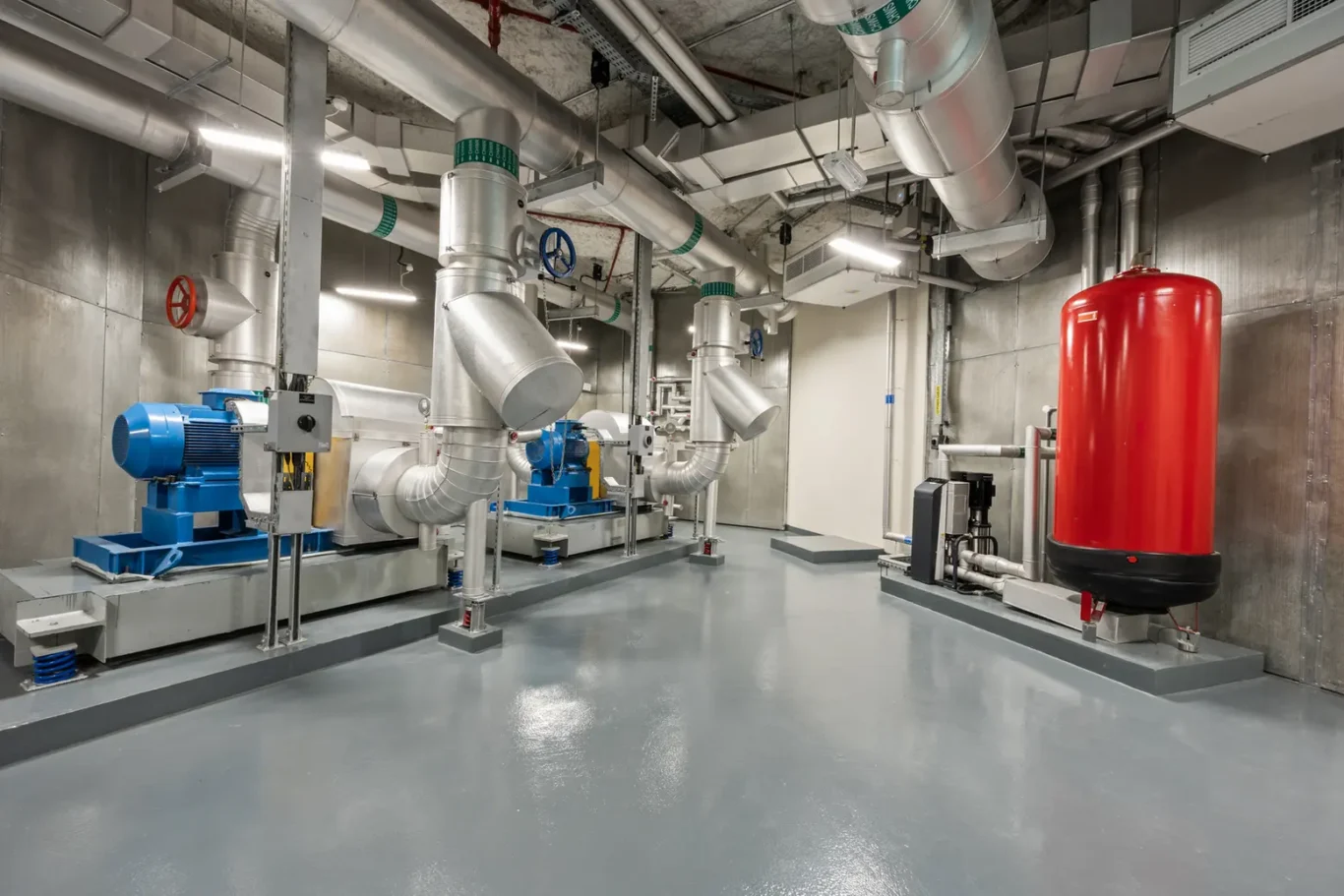 Industrial mechanical room with large metallic ducts, blue pumps, and a tall red pressure tank.