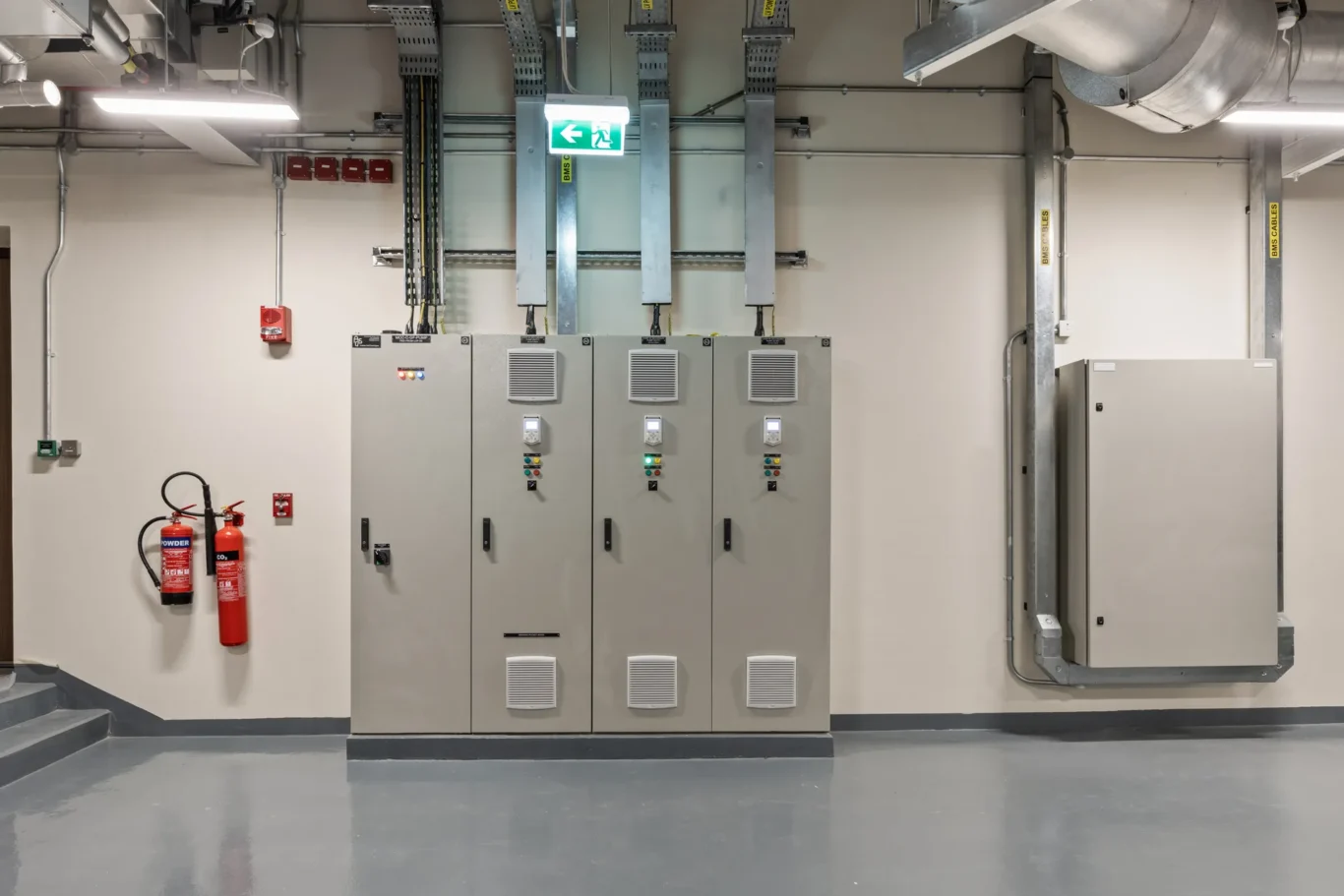 Row of gray electrical cabinets with control panels in a clean industrial room, fire extinguishers mounted on the wall nearby.