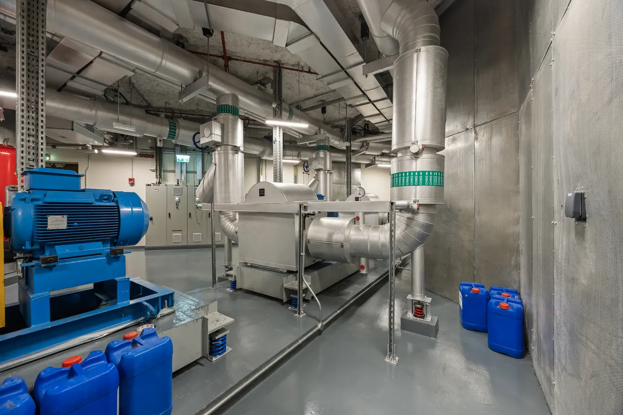 Industrial mechanical room with a large blue electric motor, extensive silver ductwork, and blue chemical containers along the wall