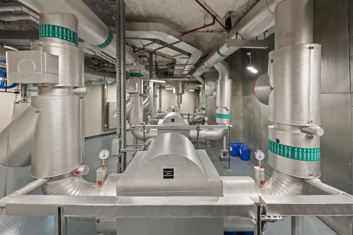 Industrial mechanical room with large silver HVAC ducts, pipes, and gauges arranged in a corridor and ceiling.]?