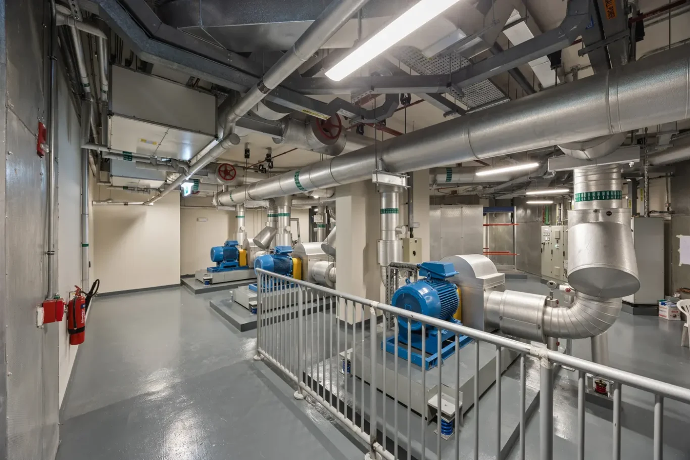 Industrial mechanical room with large silver ducts, piping, and blue motors; safety railing in foreground and equipment on concrete platforms.