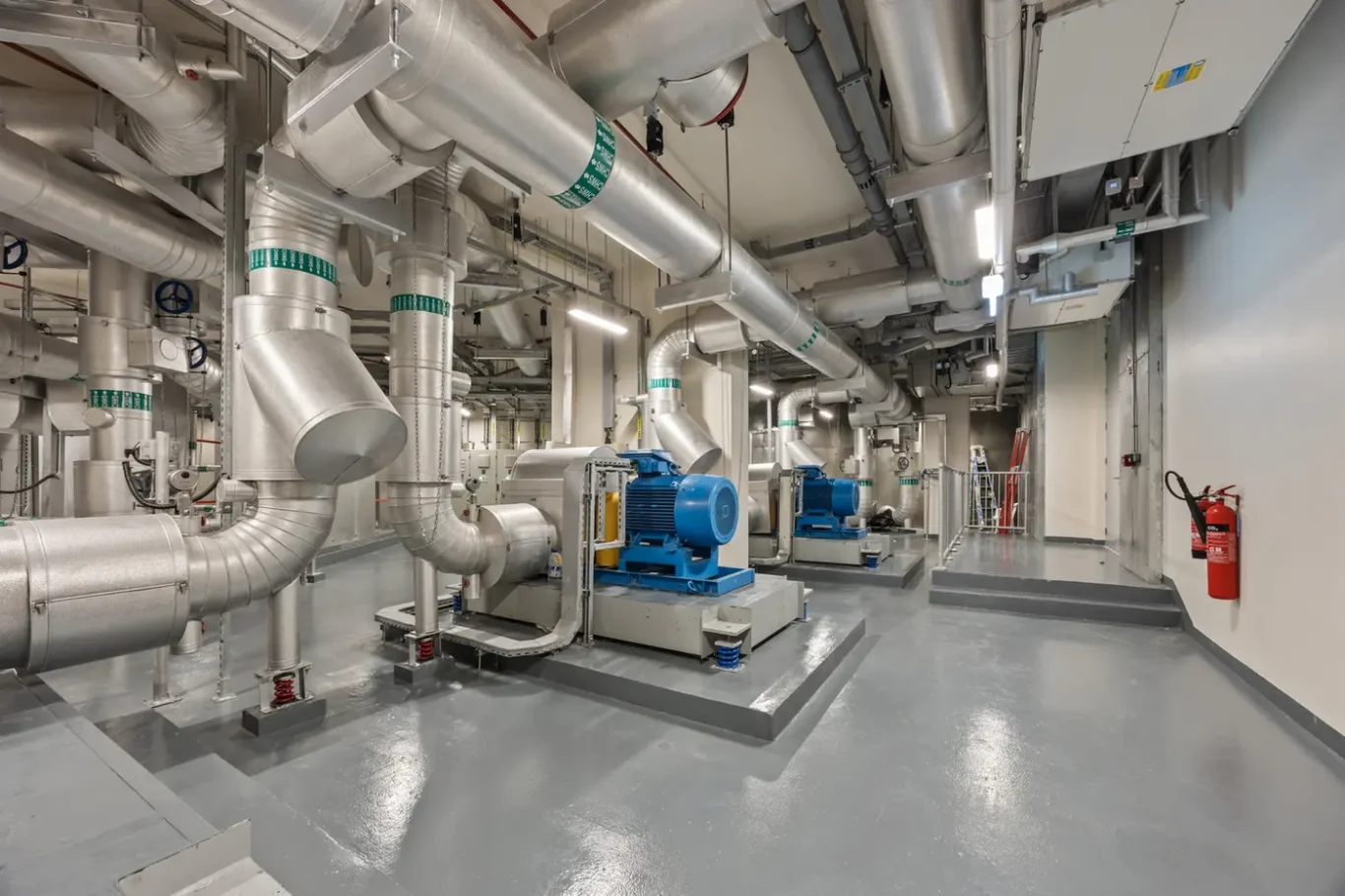 Industrial mechanical room with large silver ductwork and blue pumps on raised platforms.
