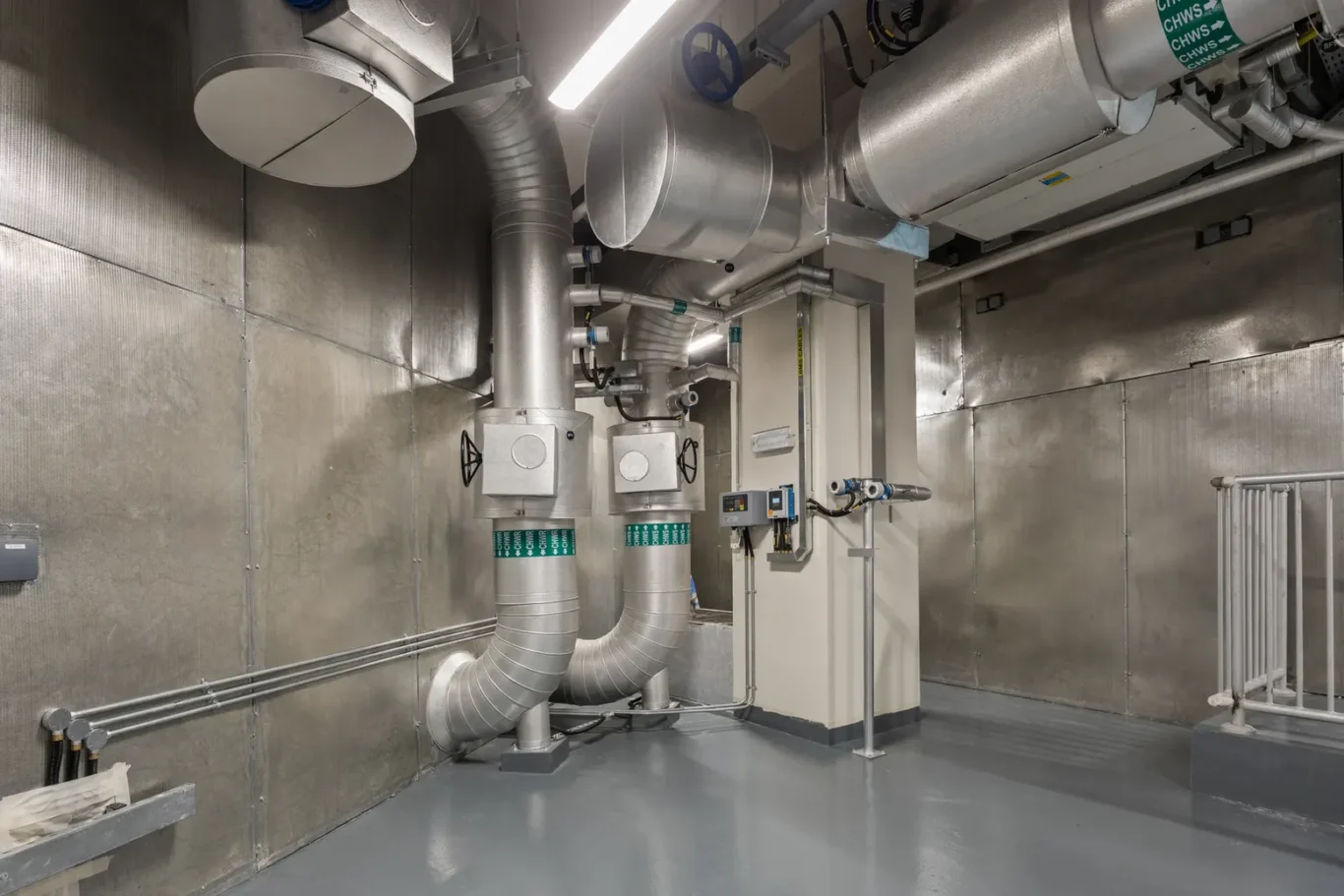 Industrial mechanical room with large silver ducts, pipes, and control panels mounted on metal walls and gray floor.