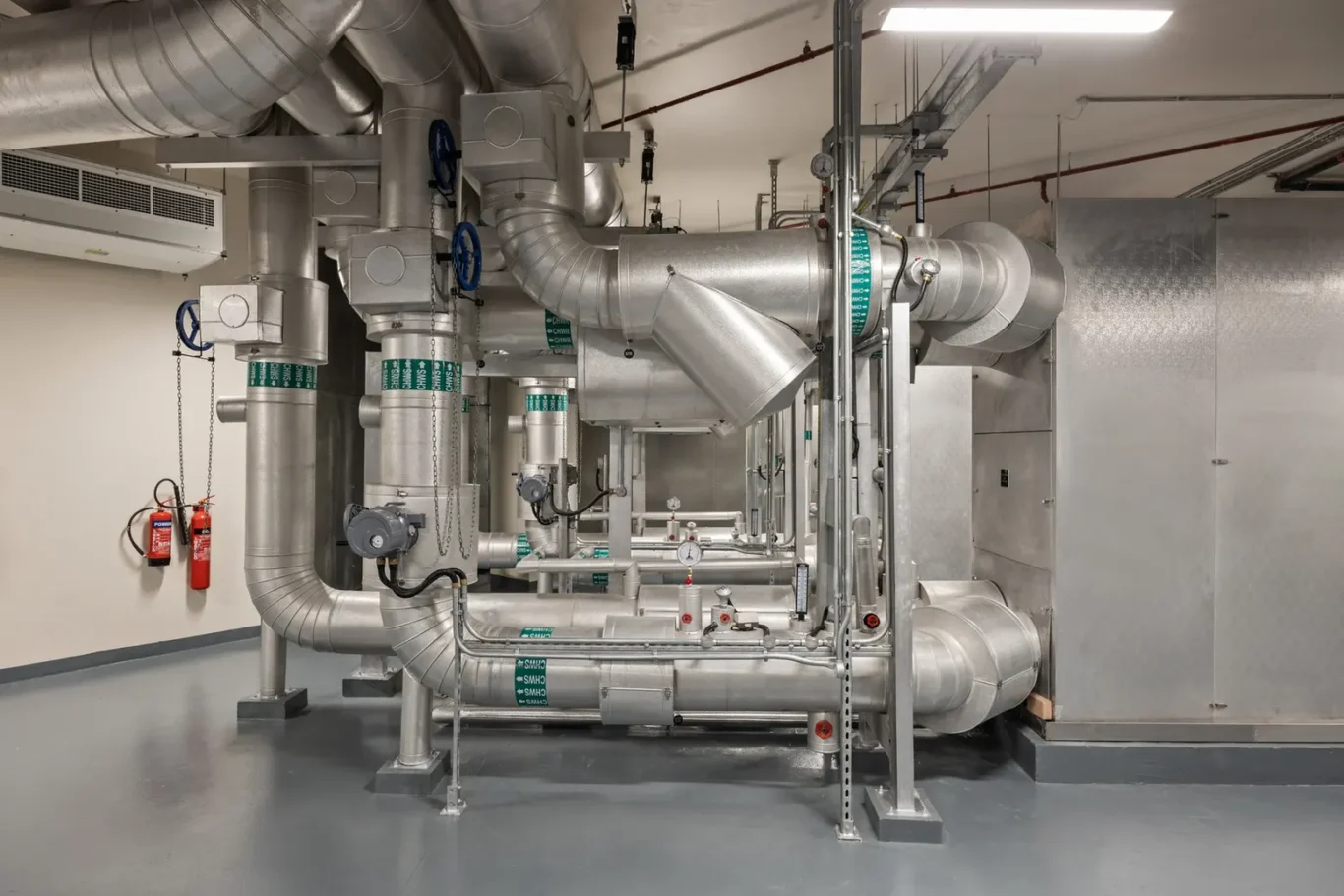 Industrial mechanical room with large silver HVAC ducts, pipes, and valves; red fire extinguishers mounted on a wall to the left.