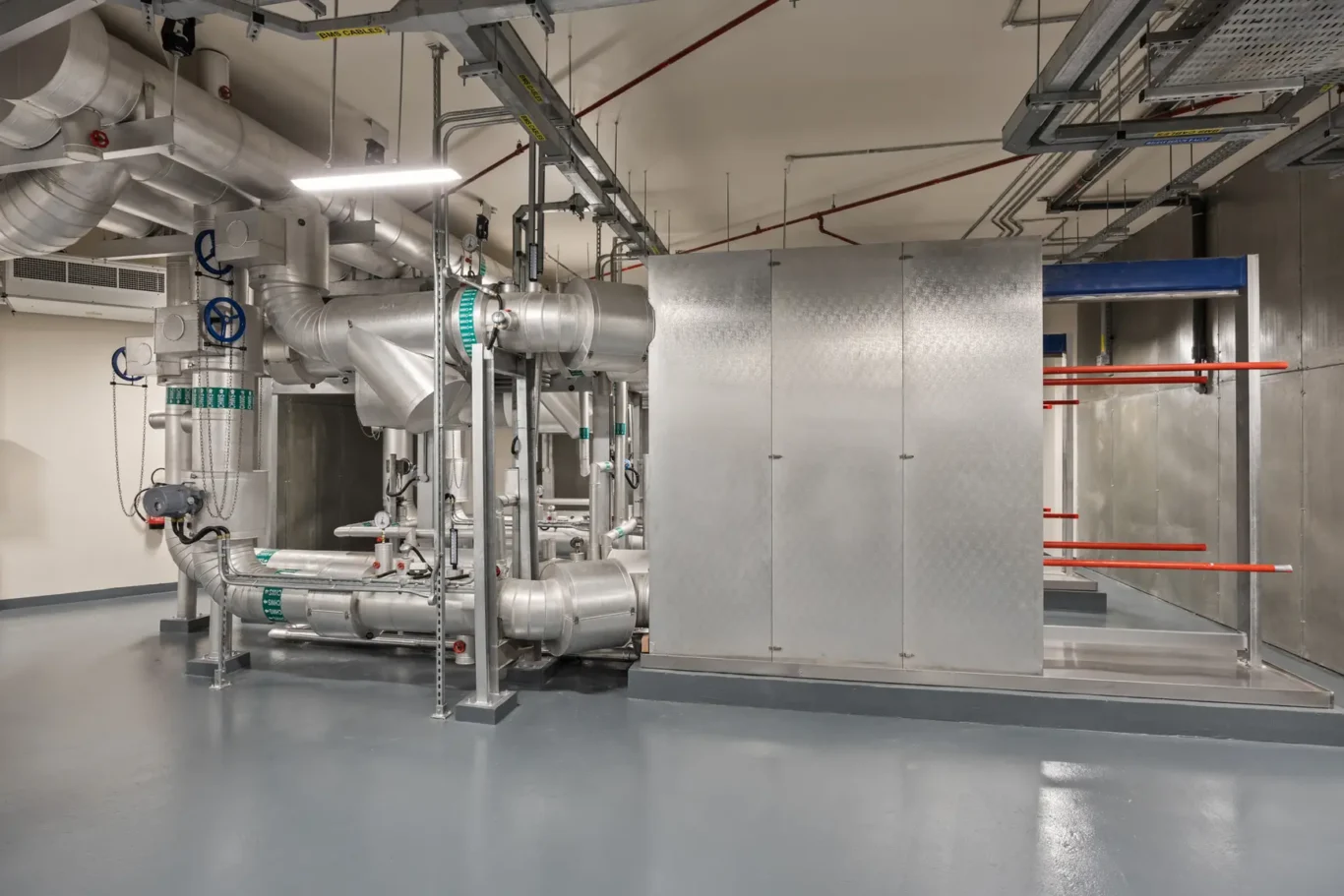 Industrial mechanical room with a dense network of stainless steel pipes, valves, and ducts, plus a tall diamond-plate metal cabinet on the right and red support pipes along the wall.