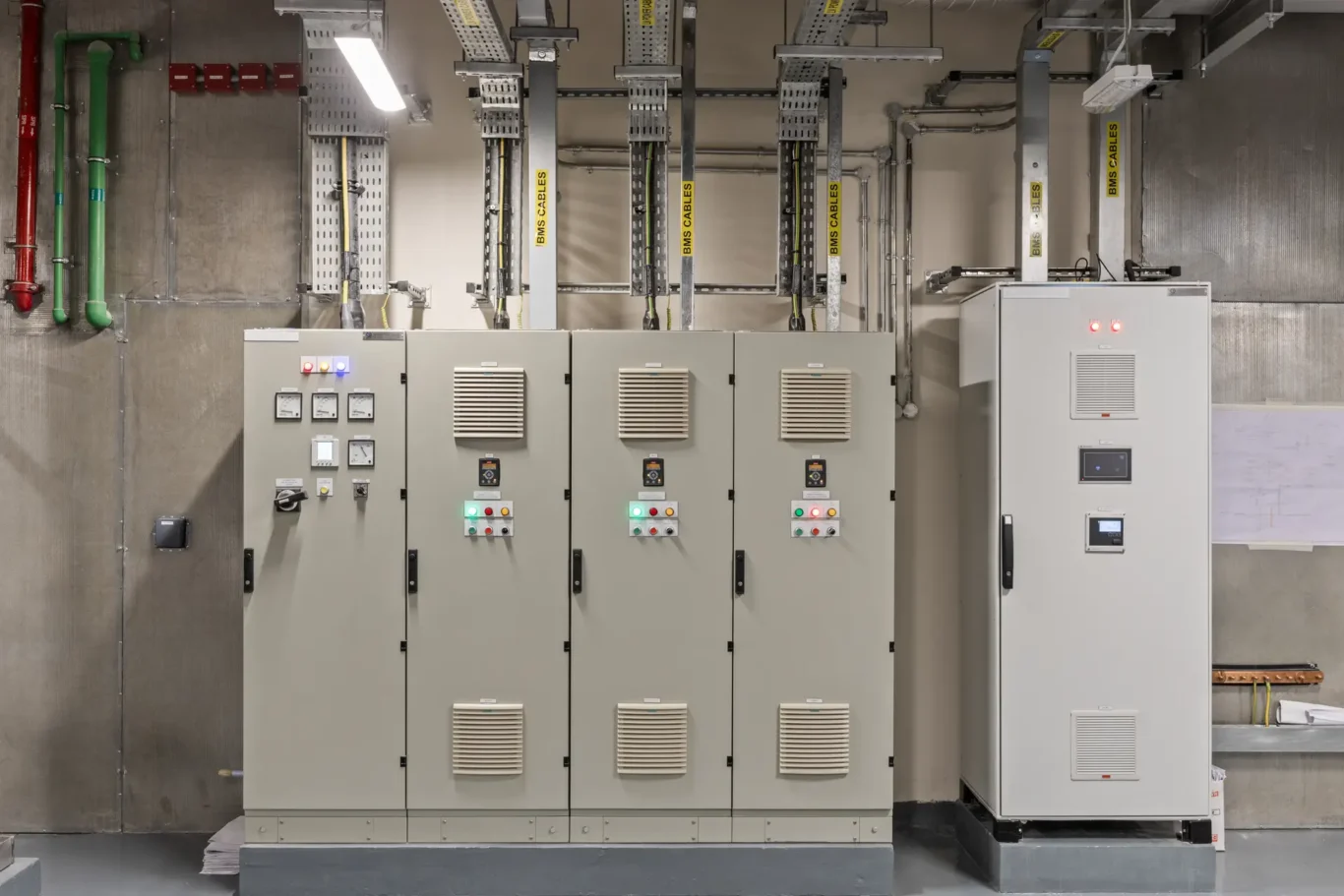 Industrial electrical control room with a row of grey control panels, gauges, and push buttons. Overhead cable ladders labeled 'BMS CABLES' connect to the cabinets.