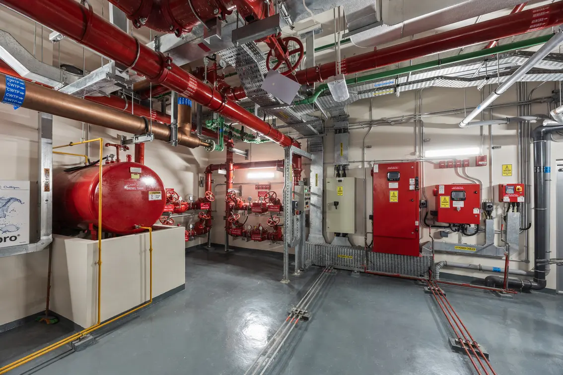 Industrial mechanical room with red pipes, valves, and control panels on the wall.