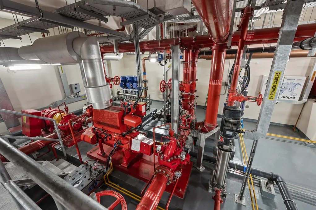 Industrial mechanical room with red piping, valves, and a large red pump assembly in the foreground.