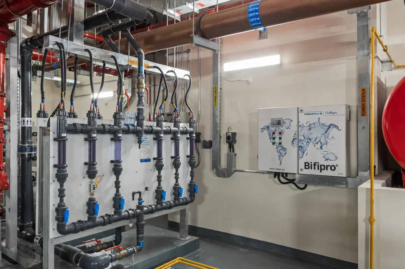 Industrial mechanical room with a row of vertical pipes, valves, and fittings connected to a metal frame. Red and copper pipes run overhead with blue-handled valves at each line amplifying a complex piping system.
