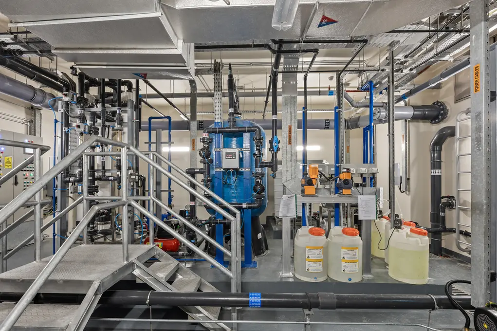 Industrial equipment room with blue processing tank, network of pipes, valves, and metal stairs.