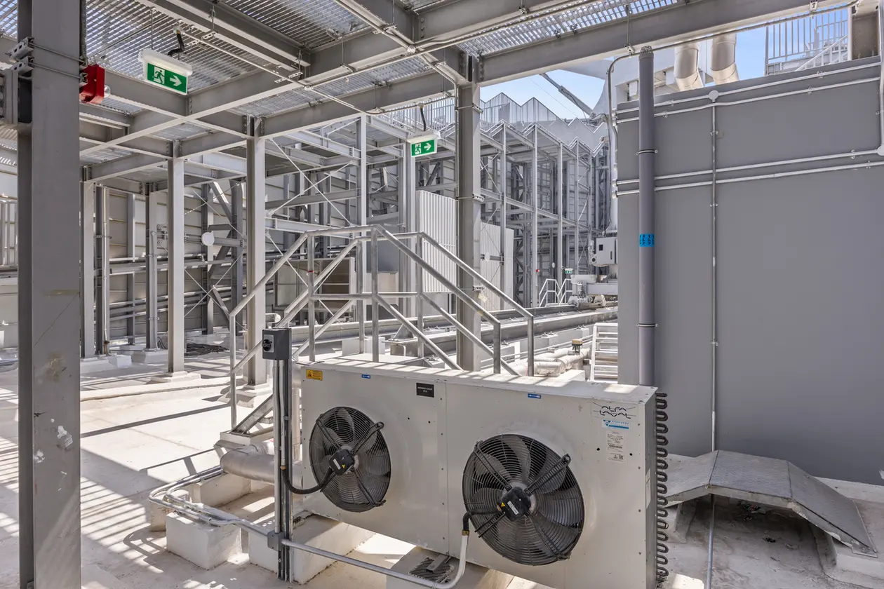 Industrial mechanical area with metal walkways, staircases, and large cooling unit with two fans; exposed pipes and framework overhead.