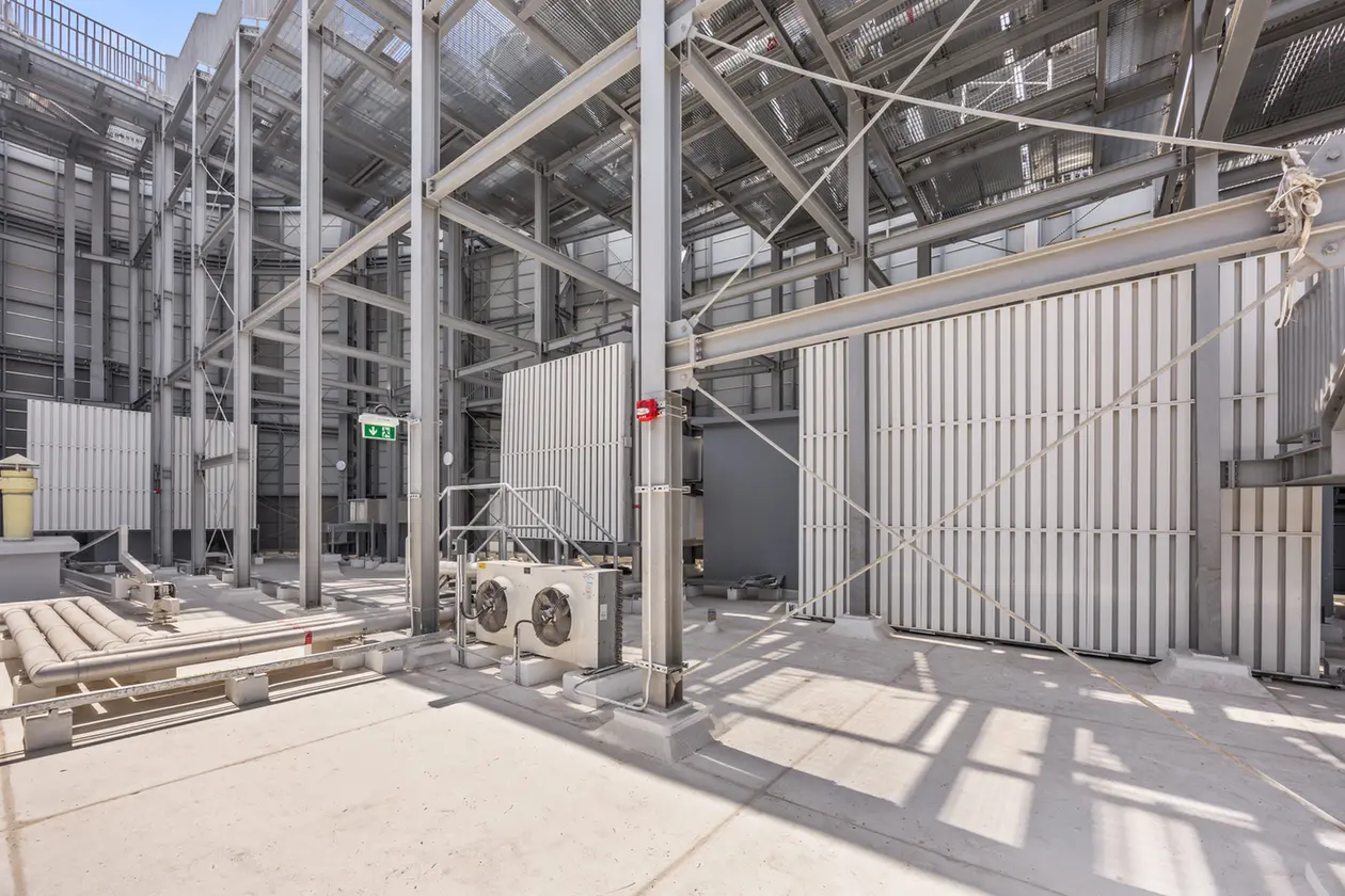 Industrial facility interior with steel framework, pipes, and ventilation panels; a green exit sign visible in the background.