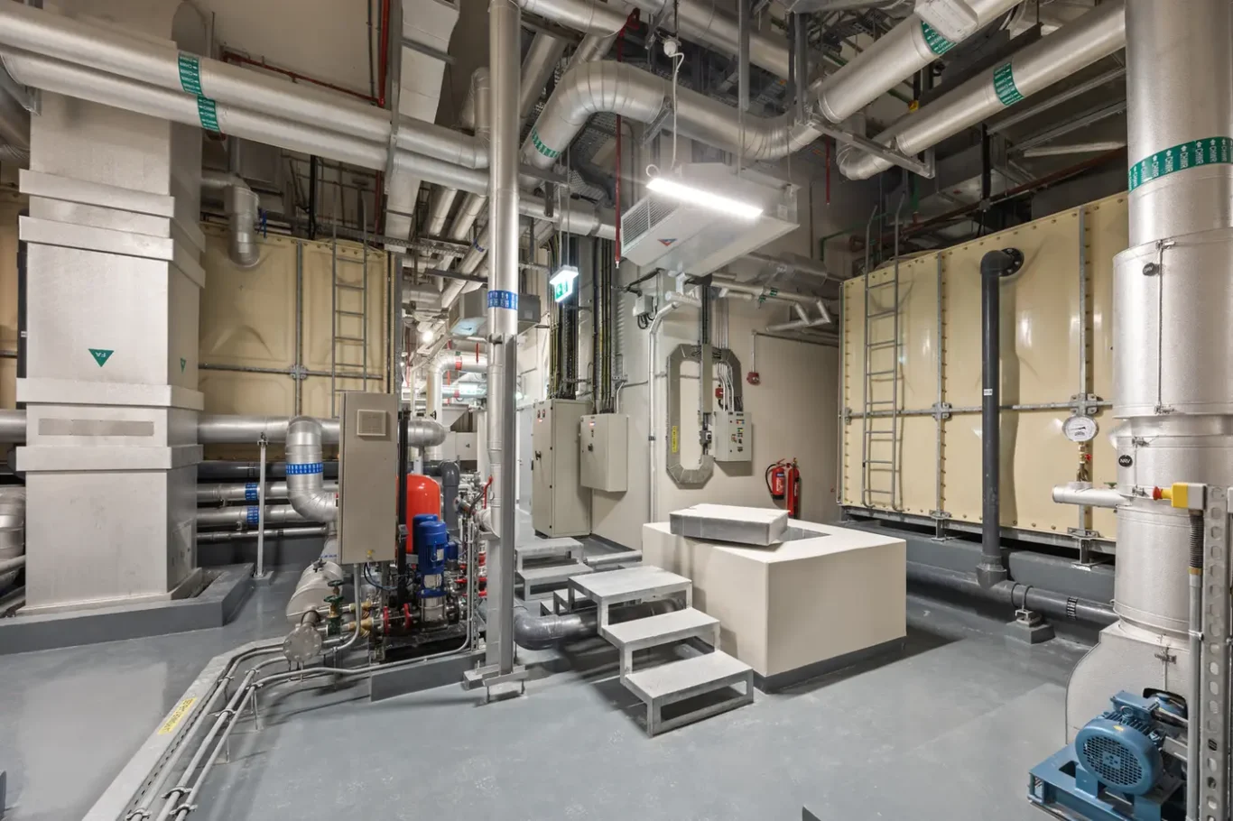 Industrial mechanical room with large metal pipes, ducts, and equipment including pumps and control panels.
