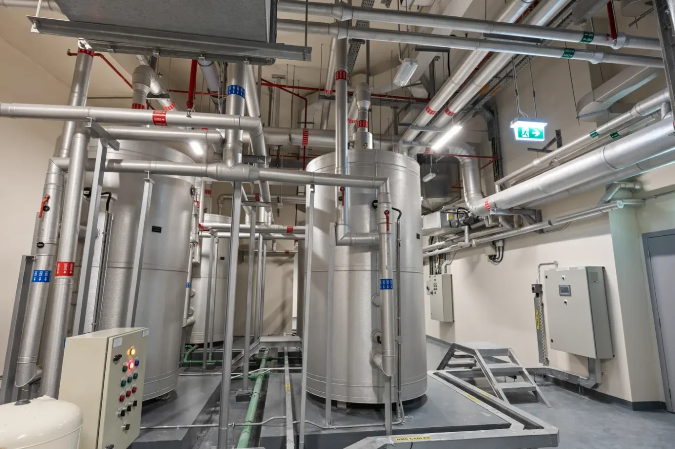 Industrial mechanical room with large stainless steel tanks, a network of pipes, valves, and a control panel to the left.