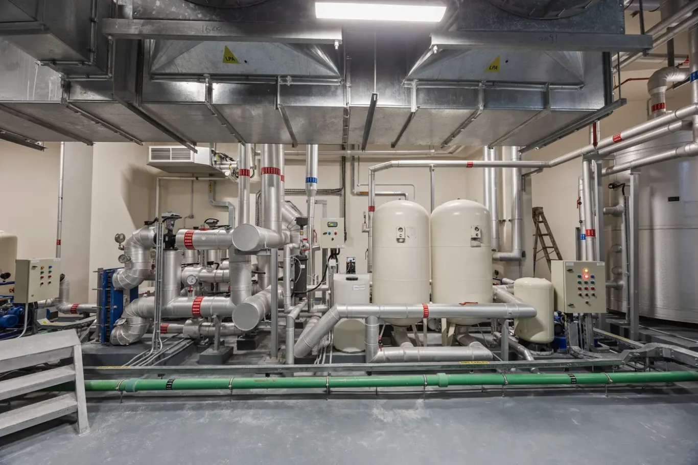 Industrial mechanical room with a dense network of silver pipes, gauges, and two large white tanks/pressure vessels in the center.