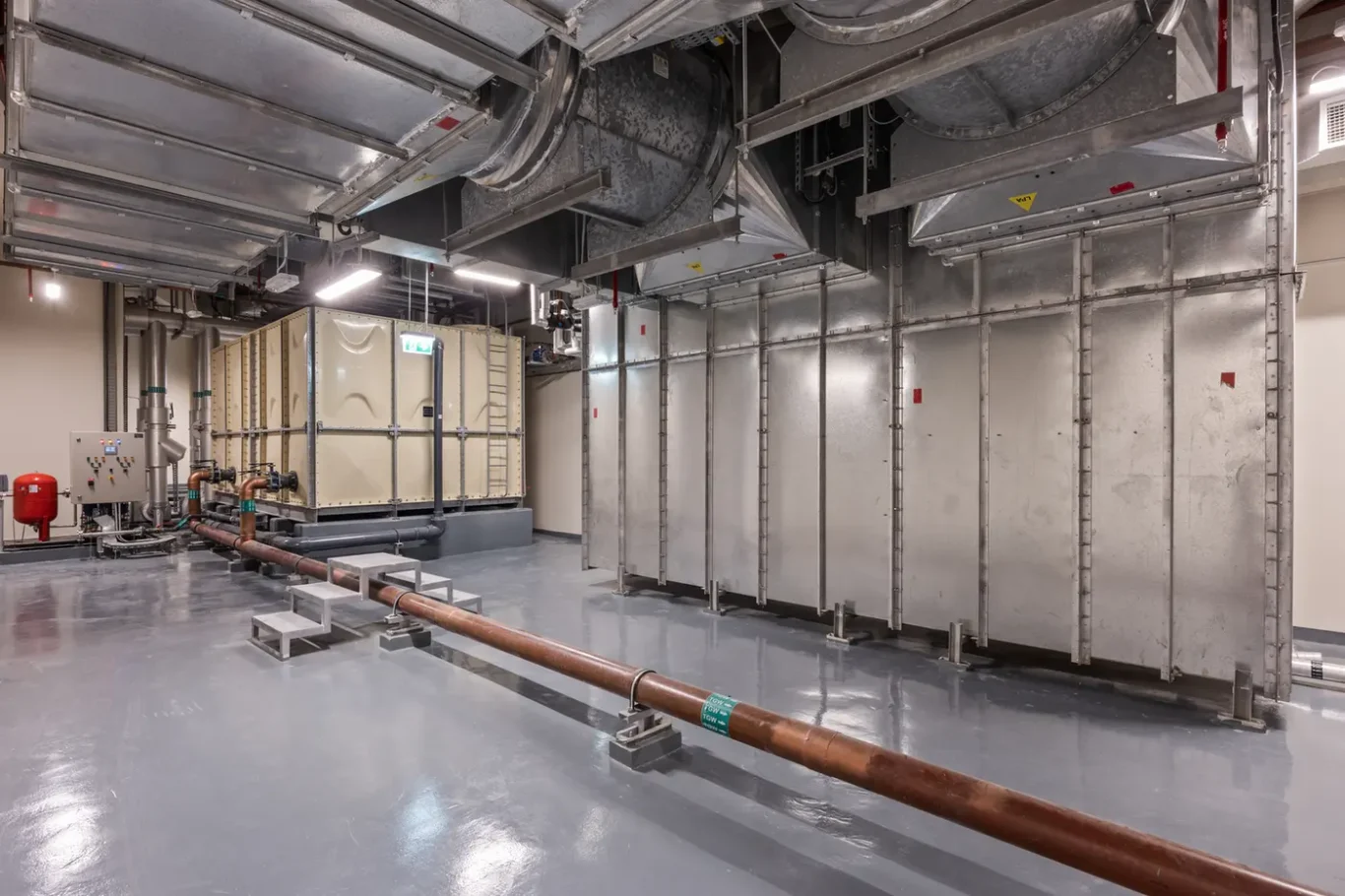 Industrial mechanical room with large galvanized HVAC ducts, metal panels, and exposed piping; copper pipe runs along the floor on supports with a control panel and red extinguisher to the left.