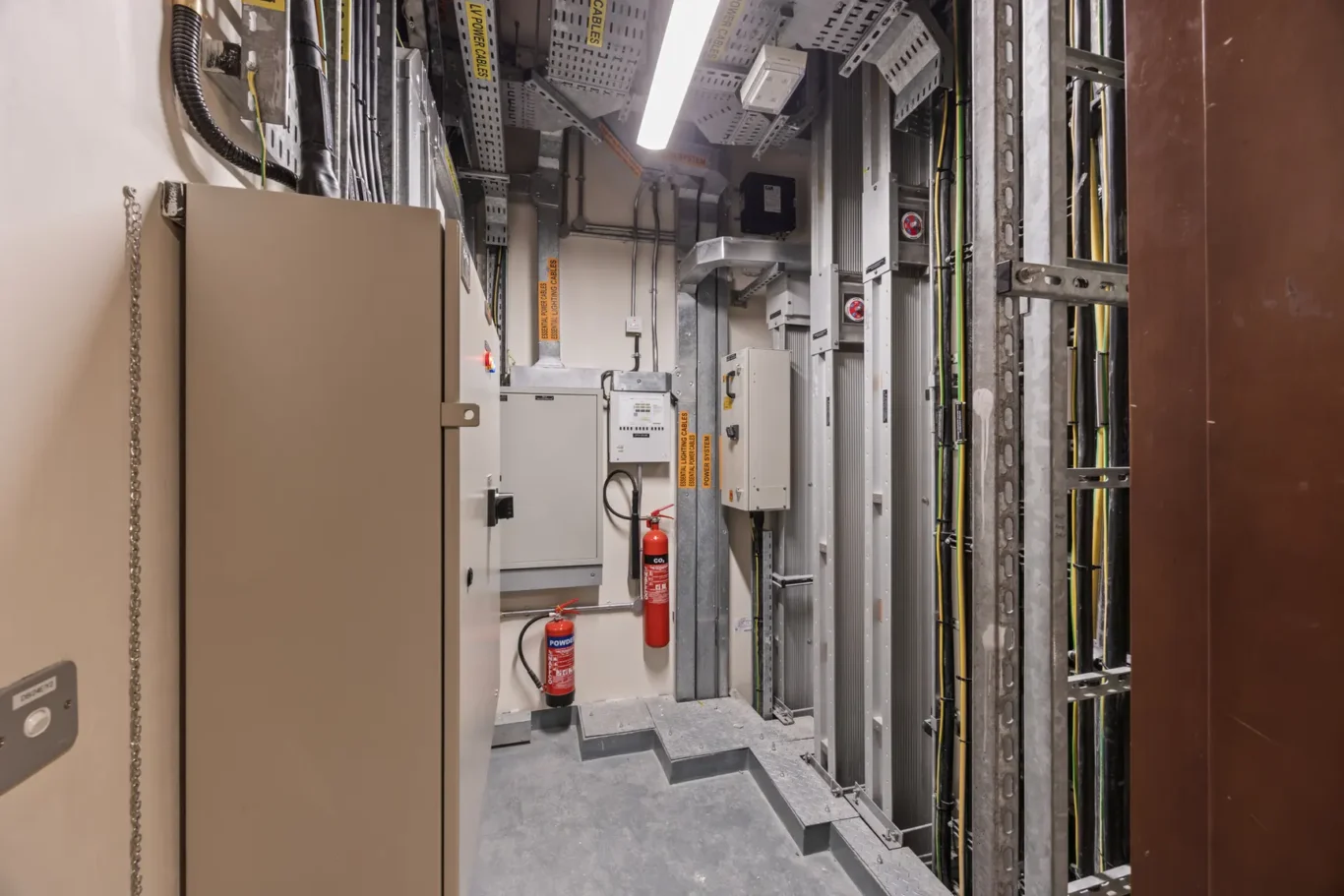 Industrial electrical equipment room with control panels, metal racks, and exposed cables. A red fire extinguisher sits near the wall on the left.