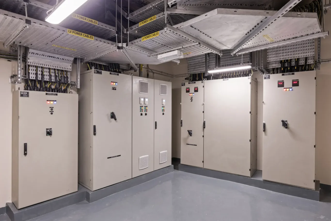 Row of electrical switchgear and beige cabinets in a power distribution room, with overhead cable trays labeled 'LV POWER CABLES'.