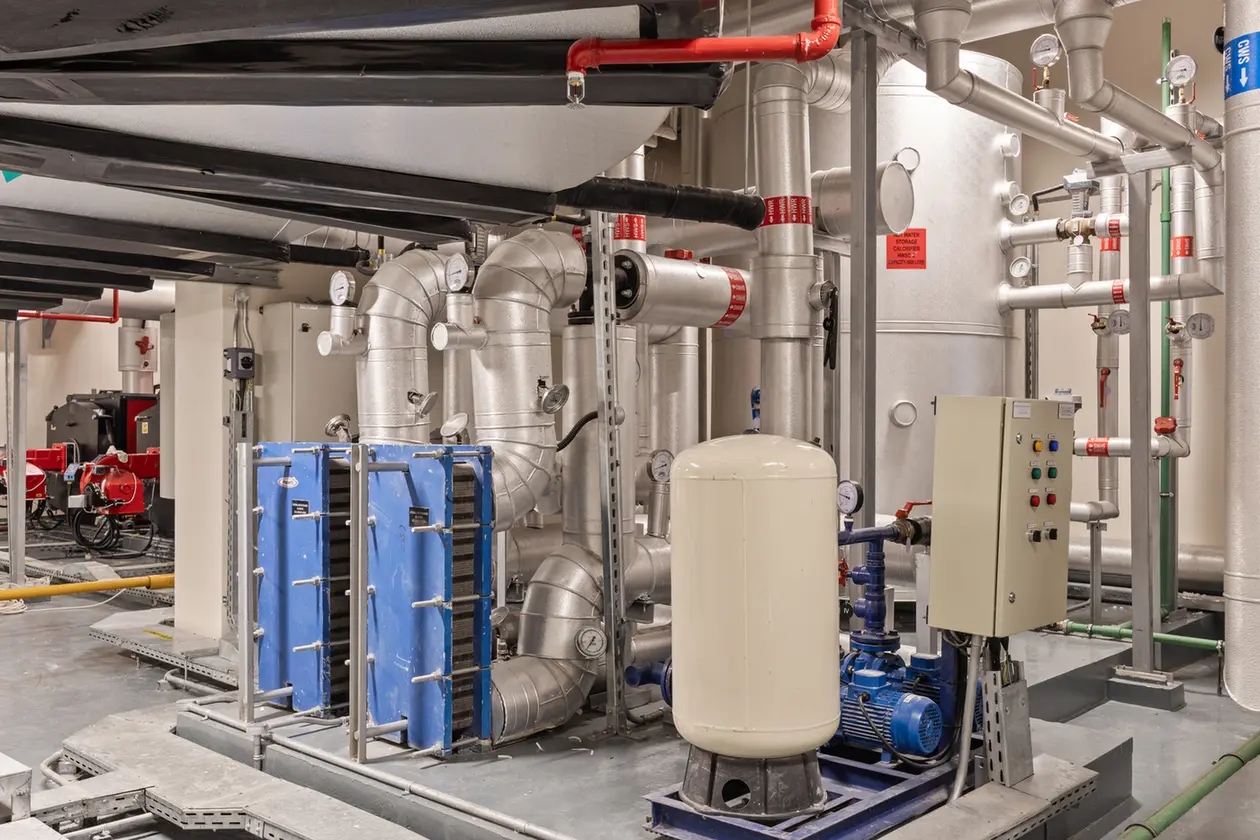 Industrial mechanical room with interconnected insulated pipes, gauges, a control panel, and a white pressure tank on a blue base.