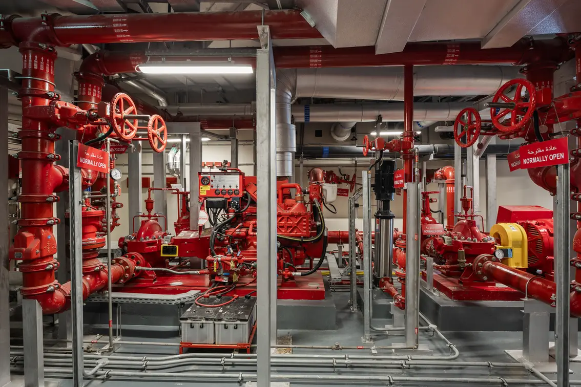 Industrial mechanical room with red fire-suppression piping, valves, and pumps; signs indicate 'Normally Open.'