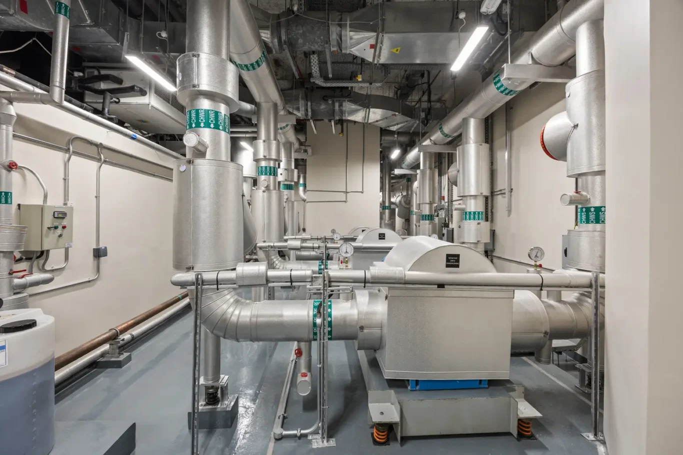 Industrial mechanical room with a dense network of silver insulated pipes, ducts, and valves surrounding a central machine assembly.