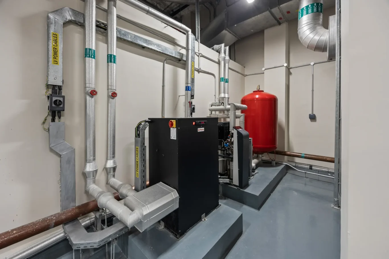 Industrial boiler room featuring a black furnace, silver insulated pipes, and a red expansion tank against beige walls.
